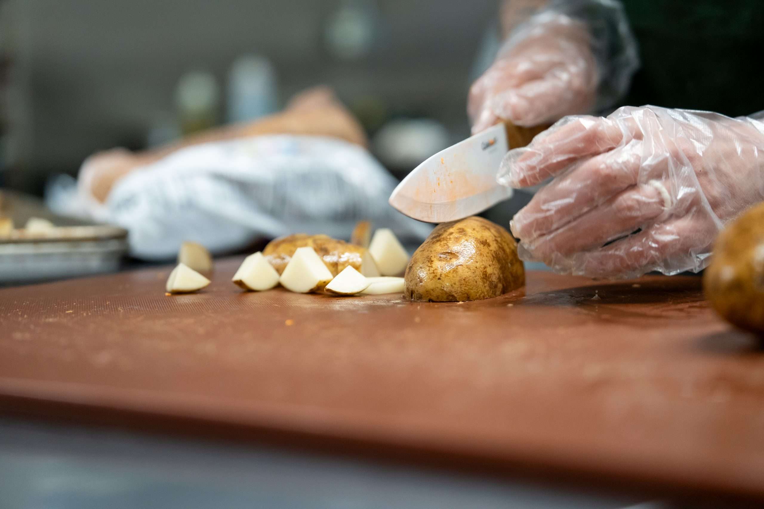 Chef wearing clear plastic gloves slicing golden potatoes on wooden cutting board in professional kitchen