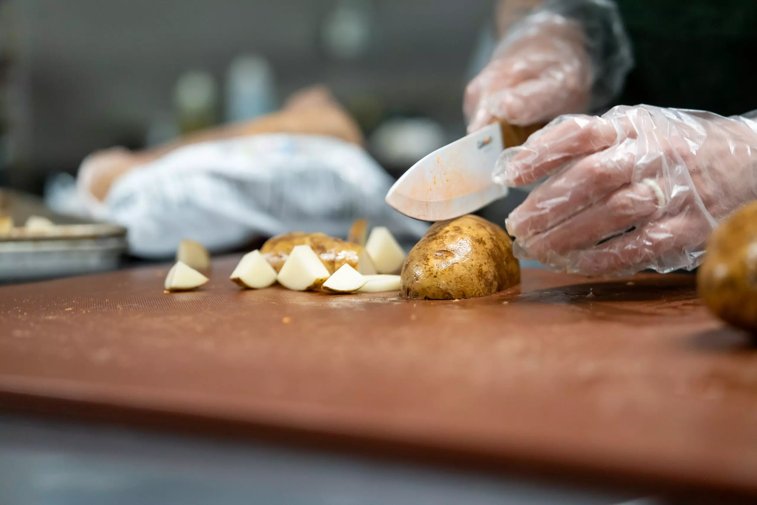 Chef wearing clear plastic gloves slicing golden potatoes on wooden cutting board in professional kitchen