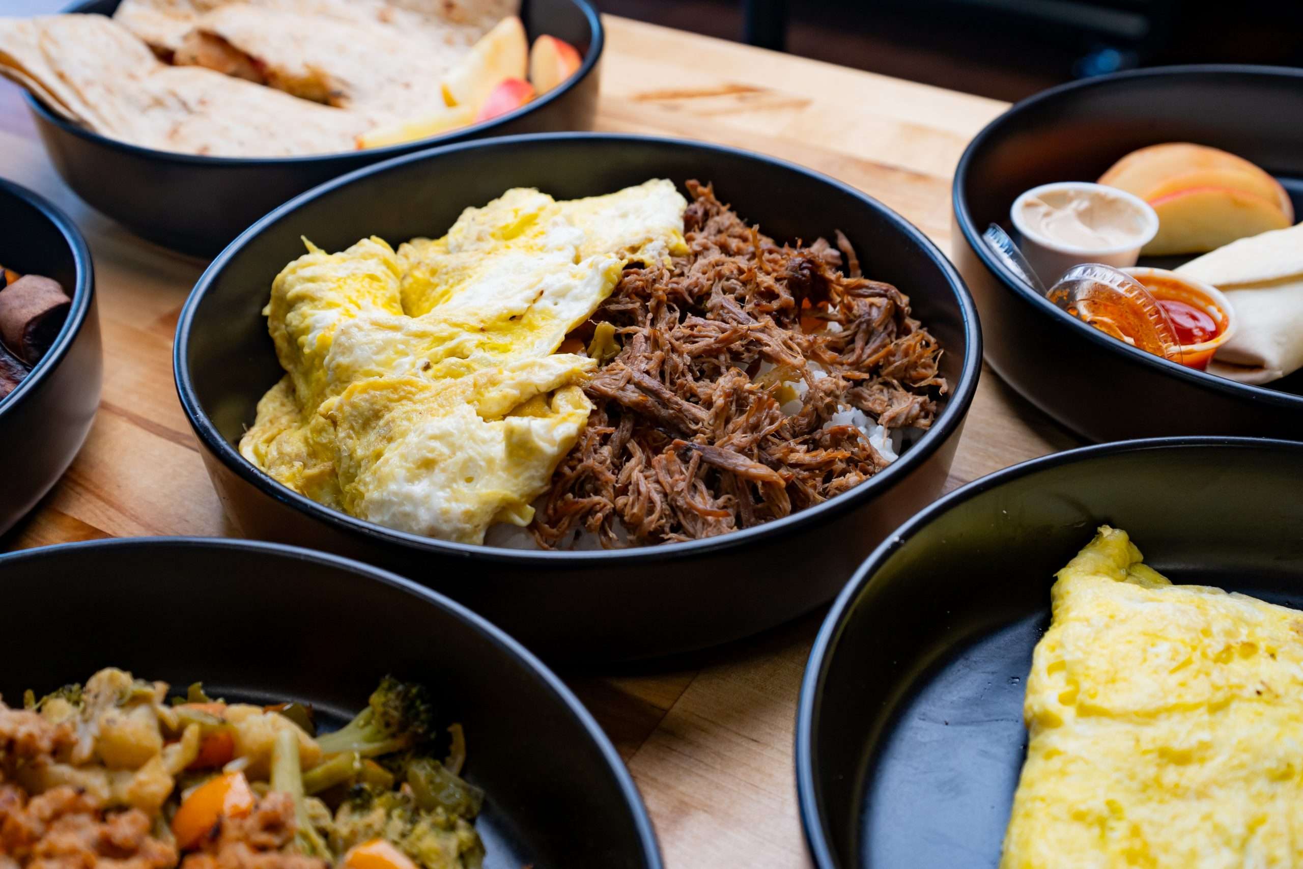 Multiple black bowls with scrambled eggs, shredded beef, vegetables, and condiments arranged on wooden table